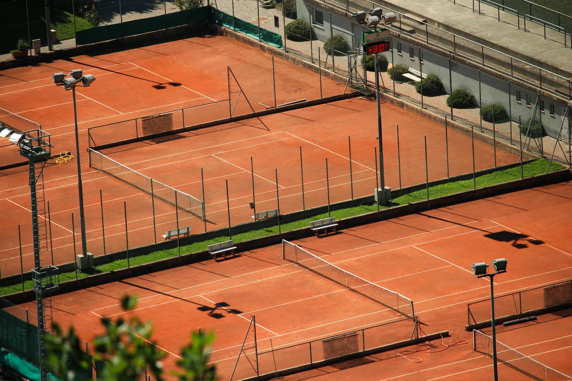 Aerial shot of empty red clay tennis courts on a sunny day with shadows.