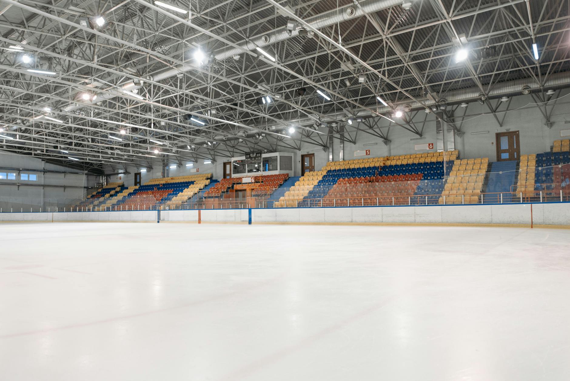 An empty indoor ice rink with colorful seats, bright overhead lights, and a clear ice surface.