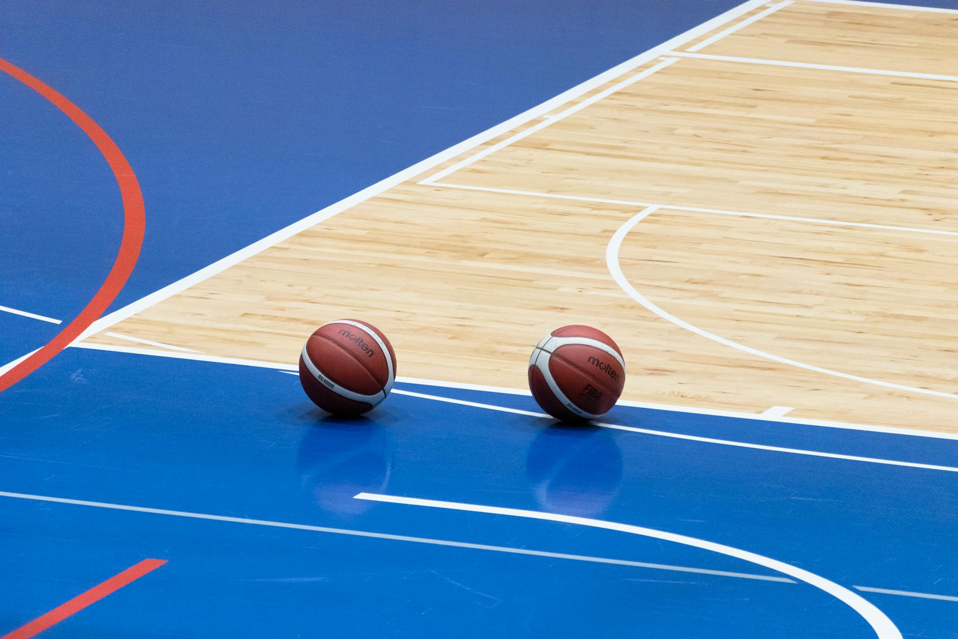 Two basketballs resting on a blue indoor basketball court with wooden flooring.