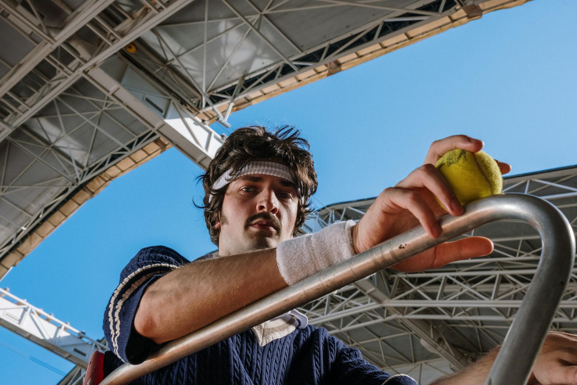 Low angle view of a tennis player holding a ball beneath a stadium roof. Bright, clear sky.